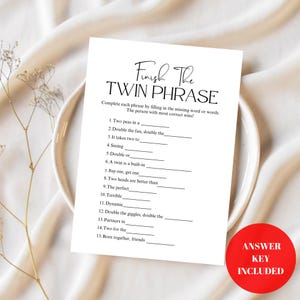 May include: A white card with the text "Finish The TWIN PHRASE" printed in black, with fill-in-the-blank phrases. A red circle with "ANSWER KEY INCLUDED" is in the lower right corner. The card is on a white plate.