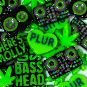 May include: Assortment of green and black resin buttons with various designs, including a heart with the word "PLUR", a marijuana leaf, a boombox, and the words "Bass Head" and "Where's Molly".