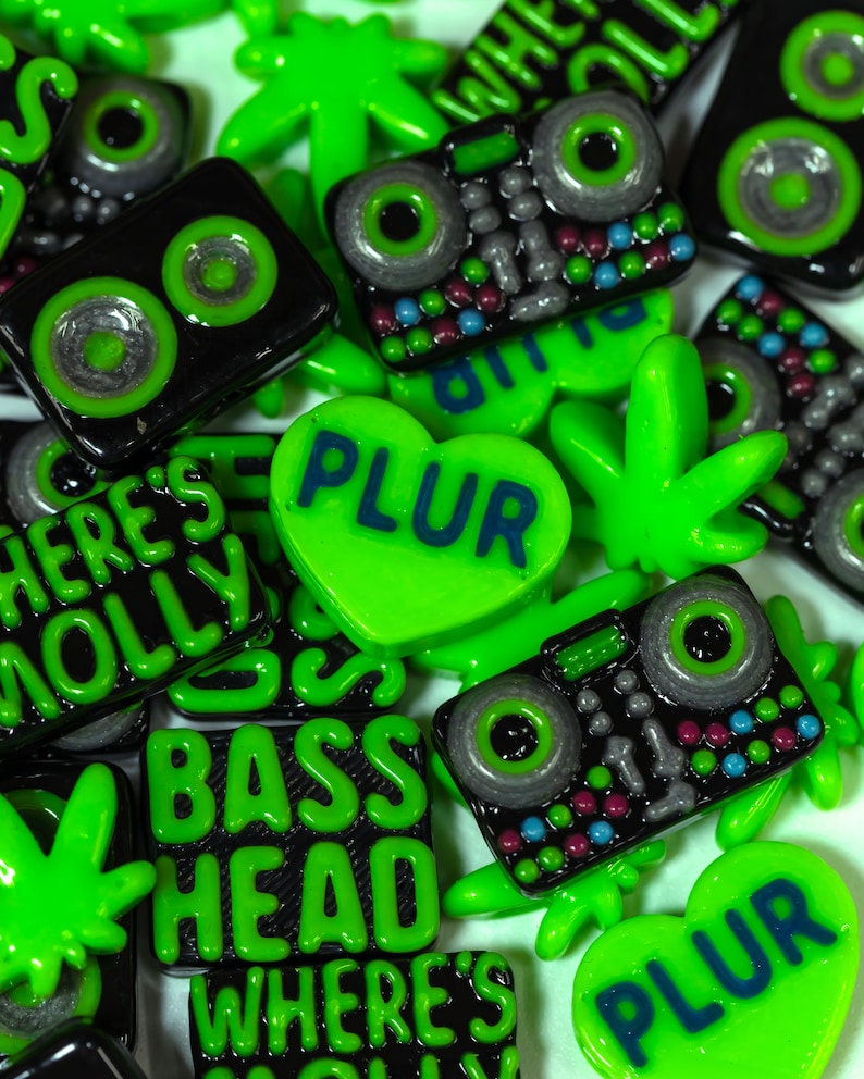 May include: Assortment of green and black resin buttons with various designs, including a heart with the word "PLUR", a marijuana leaf, a boombox, and the words "Bass Head" and "Where's Molly".
