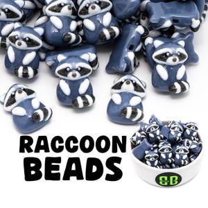 May include: A collection of blue and white raccoon-shaped beads. The beads have black and white striped tails and are arranged in a bowl and scattered. The text "RACCOON BEADS" is displayed in bold, black letters.