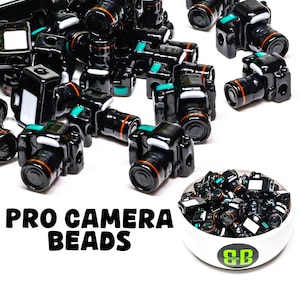 May include: Black camera beads with orange and teal accents are scattered across a white surface. The text "PRO CAMERA BEADS" is displayed in bold, black letters. A small bowl filled with similar beads is also present.