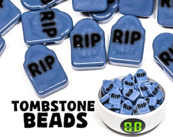 May include: A bowl filled with blue tombstone beads with the word "RIP" printed on each bead. The beads are scattered around the bowl on a white surface. The text "TOMBSTONE BEADS" is printed below the bowl.