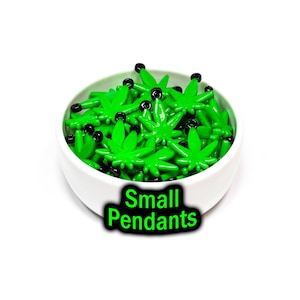 May include: A white bowl filled with small green marijuana leaf-shaped pendants with black jump rings. The bowl is sitting on a white surface. The text "Small Pendants" is written in black letters with a green outline below the bowl.