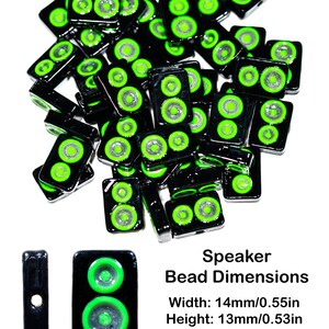 May include: A pile of black and green speaker-shaped glass beads. Each bead has two green circles with a silver center. The beads are 14mm wide and 13mm high with a 2mm hole.