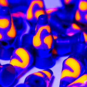Może przedstawiać: Kolekcja małych lamp lawowych o retro designie. Lampy mają niebieski wygląd zewnętrzny z żywymi pomarańczowymi i ż&oacute;łtymi wzorami lawy wewnątrz. Lampy są ułożone blisko siebie, tworząc efektowną wizualnie ekspozycję.
