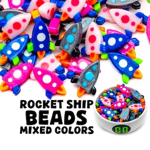 Może przedstawiać: Kolekcja kolorowych koralików w kształcie rakiety w różnych kolorach, w tym różowym, niebieskim, szarym i białym. Koraliki mają białe kropki i są idealne do rękodzieła.