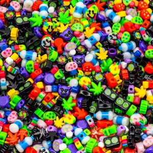 May include: A vibrant assortment of colorful plastic beads in various shapes. The beads include smiley faces, mushrooms, marijuana leaves, and other novelty designs. The color palette is diverse, featuring red, green, blue, yellow, and purple.
