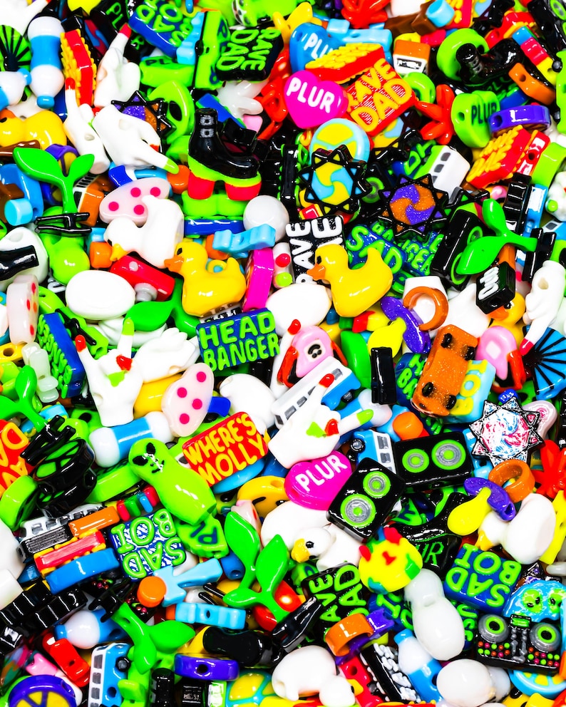 May include: A colorful assortment of plastic beads with various designs and phrases, including "PLUR", "RAVE", "SAD BOI", "HEAD BANGER", and "WHERE'S MOLLY".
