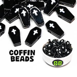 May include: Black coffin-shaped beads with white crosses are scattered across a white surface. A small white bowl filled with similar beads is on the right. The words "COFFIN BEADS" are in bold black letters.
