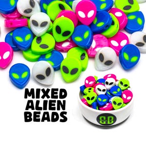 May include: A collection of colorful alien-shaped beads in green, blue, pink, and white. The beads are scattered on a white surface and in a small white bowl with a green logo. The text "MIXED ALIEN BEADS" is printed in black on a white background.