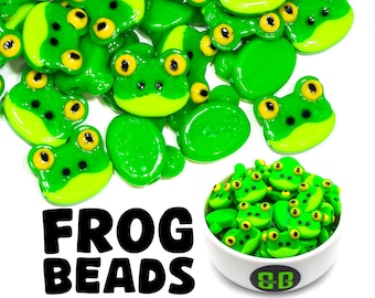 Puede incluir: Un bol lleno de cuentas verdes y amarillas con forma de rana. Las cuentas tienen ojos negros y una boca blanca. El bol es blanco con un logotipo blanco y negro que dice "SB". El texto "FROG BEADS" está escrito en letras negras sobre un fondo blanco.