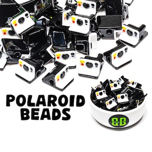 May include: A collection of black and white Polaroid camera beads with yellow and red accents. The beads are scattered across a white surface, with some contained in a small white bowl. The text "POLAROID BEADS" is displayed in large, bold letters.