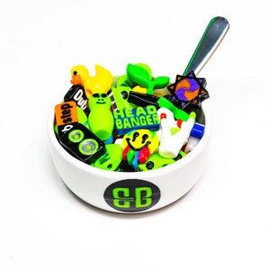 May include: A white ceramic bowl with a green "BB" logo on the bottom, filled with colorful plastic charms. The charms include a yellow duck, green aliens, a green plant, a smiley face, a speaker, and a charm that says "Head Banger".