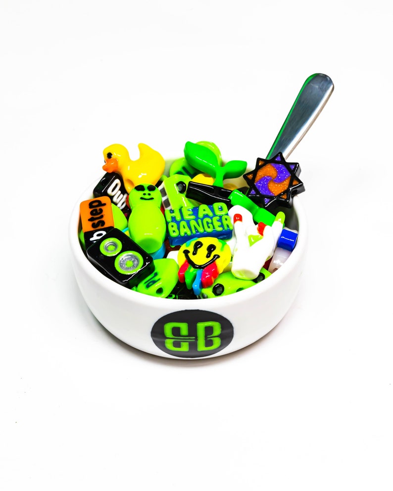May include: A white ceramic bowl with a green "BB" logo on the bottom, filled with colorful plastic charms. The charms include a yellow duck, green aliens, a green plant, a smiley face, a speaker, and a charm that says "Head Banger".