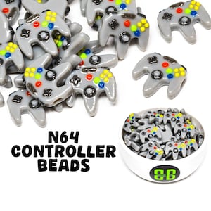May include: Gray N64 controller beads with colorful buttons are scattered across a white surface. A white bowl filled with the beads is in the lower right corner. The text "N64 CONTROLLER BEADS" is in black letters.