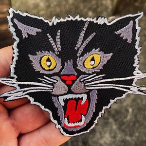 May include: Black embroidered iron-on patch of a snarling cat with yellow eyes and a red mouth.