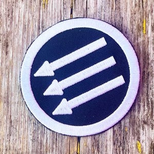 Può includere: Toppa rotonda con sfondo blu navy e tre frecce bianche rivolte verso il basso e verso destra. Le frecce sono all'interno di un bordo circolare bianco. La toppa è su una superficie di legno usurata.