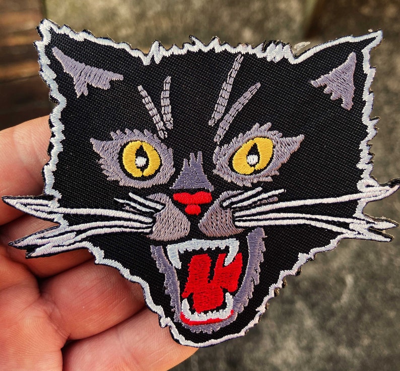 May include: Black embroidered iron-on patch of a snarling cat with yellow eyes and a red mouth.