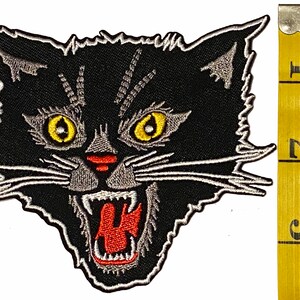 May include: Black and white embroidered patch of a snarling black cat's head with yellow eyes and red mouth. The patch is approximately 5 cm wide.