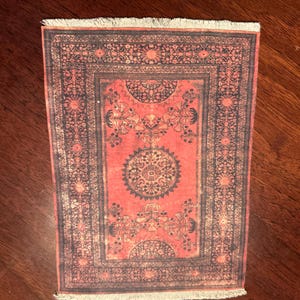 May include: A small rectangular rug featuring a red field with detailed dark blue floral patterns. The rug has a central medallion design and a fringed edge, reflecting a traditional oriental style.