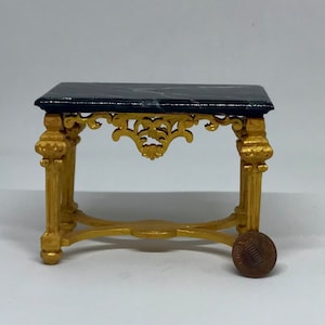 May include: Miniature black marble-topped table with ornate gold-colored legs and decorative accents. The table is approximately 3 inches long and 2 inches wide, with a detailed, vintage design. A penny is next to the table for scale.