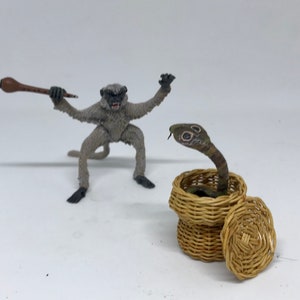 May include: A gray monkey figurine holding a wooden flute, facing a brown wicker basket with a lid. A green and brown snake figurine is inside the basket with its head sticking out.