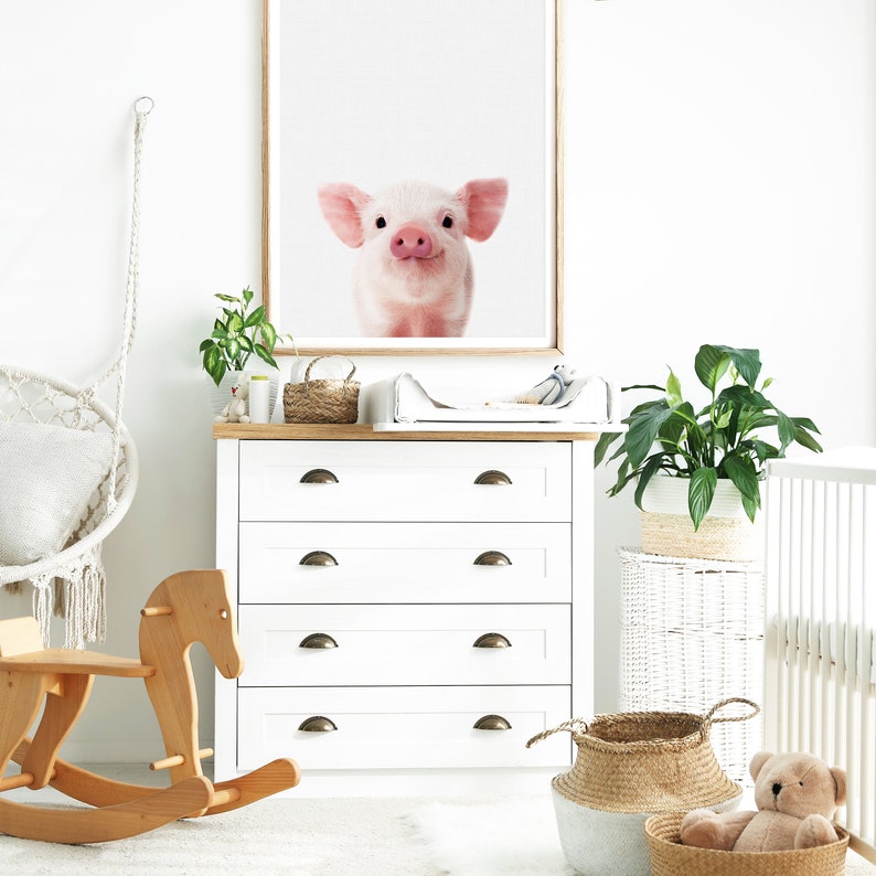 May include: A white dresser with four drawers and gold handles. A white changing table with a white changing pad is on top of the dresser. A rocking horse is on the left side of the dresser. A potted plant is on the right side of the dresser. A wicker basket is on the floor next to the dresser. A teddy bear is sitting in the wicker basket. A white crib is in the background.