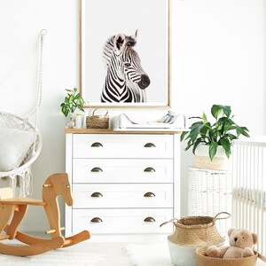 May include: A white dresser with four drawers and brass handles. A rocking horse, a framed print of a zebra, a potted plant, and a wicker basket are in the room.