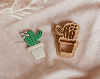 May include: Two cactus-shaped cookie cutters, one green and one brown, on a light pink fabric background. The green cutter has a pink flower on top and is in a white pot. The brown cutter is a more abstract representation of a cactus.