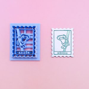 May include: A light blue plastic cookie cutter shaped like a postage stamp featuring a lizard climbing a branch and the word MÚSICA, alongside a white stamped impression of the same design with a green outline and green text on a pink background.