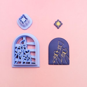 May include: A light blue plastic cookie cutter shaped like an arched window with three figures and a separate star-shaped cutter. Next to them are a dark blue arched plaque with gold embossed figures and a small gold star-shaped piece.