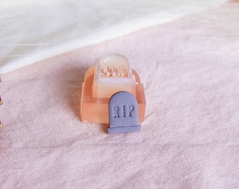 May include: A small, pink, translucent tombstone with a gray top that reads "RIP". The tombstone is sitting on a light pink fabric surface.