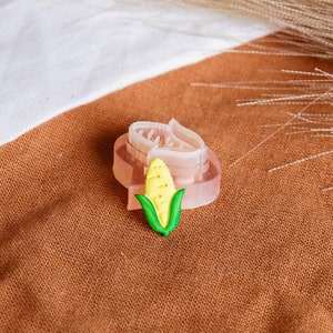 May include: A pink and clear silicone mould shaped like an ear of corn with a yellow corn cob and green leaves. The mould is sitting on a brown fabric surface.