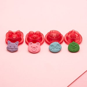 May include: Five red plastic cookie cutters shaped like animal faces, with five corresponding fondant cutouts in the colors purple, pink, blue, and green.