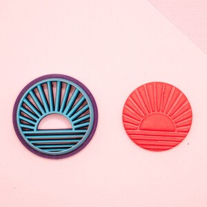 Puede incluir: Dos cortadores de galletas con forma de sol, uno azul y otro rojo, sobre un fondo rosa. El cortador azul tiene un diseño en relieve y el cortador rojo es sólido.