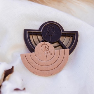 May include: A pair of geometric earrings with a layered design. The top earring is black and the bottom earring is a light brown color. Both earrings have a circular design in the center.