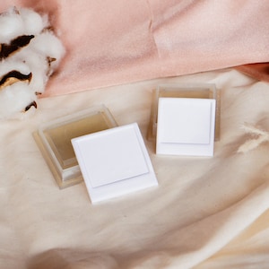 May include: Two white square containers with clear lids on a white fabric background. The containers are surrounded by cotton bolls and dried grasses.