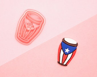 May include: Two pink plastic cookie cutters shaped like a conga drum. One cutter has a red, white, and blue Puerto Rican flag design.