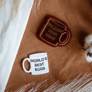 Peut inclure: Deux tasses en céramique blanche avec le texte "World's Best Boss" inscrit dessus. L'une est brune et l'autre est blanche. Les tasses sont posées sur une surface brune avec des boules de coton blanches et une plante blanche et moelleuse.