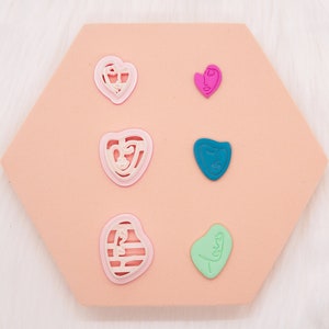 May include: Six heart-shaped resin molds with different designs. The molds are pink, white, teal, and green. They are arranged in two rows of three on a peach-colored hexagonal surface.