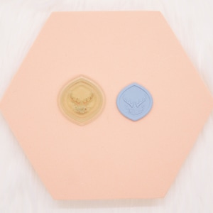 May include: Two seal stamps with a deer antler design. One stamp is clear, the other is light blue. Both are on a peach-colored, hexagonal surface. The background is white.