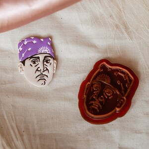 May include: Two ceramic cookie cutters shaped like a person's face wearing a purple bandana. The cookie cutters are on a white fabric background.