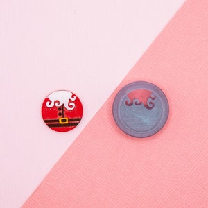 May include: Two round buttons on a pink and light pink background. One button is red with a white Santa Claus design. The other button is grey with a white design.