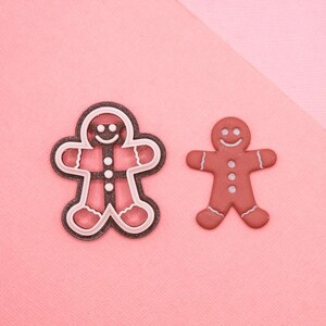 May include: Two gingerbread man decorations on a pink background. One is outlined in white with a silver interior, and the other is a smaller, solid brown color. Both have smiling faces and button details.