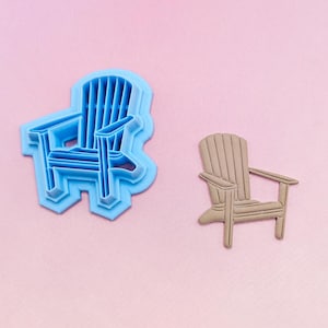 May include: A light blue plastic Adirondack chair cookie cutter positioned next to a tan-colored cutout of an Adirondack chair. The cookie cutter features a detailed outline of the chair, while the cutout shows the final shape of the pressed dough.
