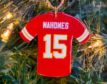 Op de afbeelding: Rode en witte voetbaltrui ornament met de naam "Mahomes" en het nummer "15" op de achterkant. De ornament hangt aan een kerstboom.