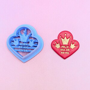 May include: A light blue heart-shaped cookie cutter and a red heart-shaped cookie with gold embossed crowns and the text FELIZ DIA DE REVES. The cutter features a mirrored design to imprint the crown graphics and text onto the red cookie surface.