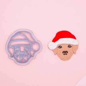 May include: A cookie cutter and a decorated cookie of a dog wearing a Santa hat. The cookie cutter is gray and the cookie is brown with white fur and a red Santa hat.