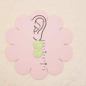 May include: A pair of green geometric earrings with a textured surface. The earrings are shown on a pink ear model with a ruler in the background.