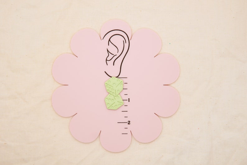 May include: A pair of green geometric earrings with a textured surface. The earrings are shown on a pink ear model with a ruler in the background.